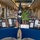A memorial display is set up on blue-draped tables in what appears to be a military base exchange or shopping corridor. The centerpiece features a POW/MIA flag alongside a small decorative tree with yellow ribbons, flanked by multiple framed informational panels containing photographs and text. A sign in the center reads 'In Memory' and 'A Promise of Remembrance,' indicating this is a tribute exhibit honoring fallen or missing service members.