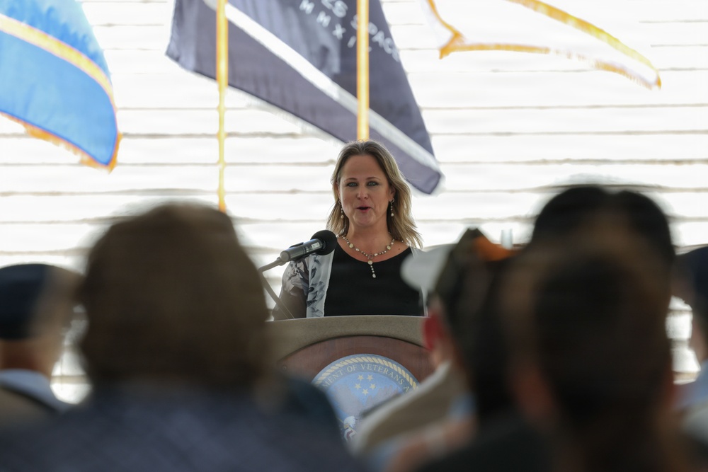 A woman with blonde hair is speaking at a podium bearing a seal that reads 'Department of Veterans Affairs.' She is addressing an audience seated in front of her, with several flags visible in the background including what appears to be a POW/MIA flag. The setting appears to be an official Veterans Affairs ceremony or event.