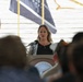A woman with blonde hair is speaking at a podium bearing a seal that reads 'Department of Veterans Affairs.' She is addressing an audience seated in front of her, with several flags visible in the background including what appears to be a POW/MIA flag. The setting appears to be an official Veterans Affairs ceremony or event.