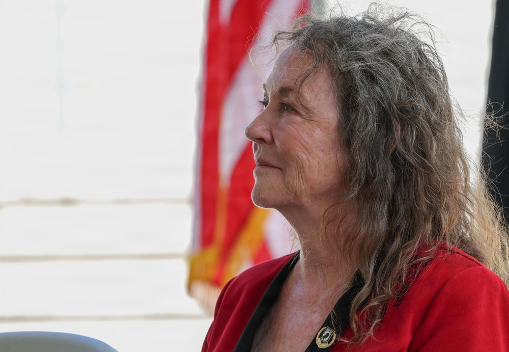 A middle-aged to older woman is photographed in profile view, gazing upward or into the distance. She is wearing a red jacket with a small pin or brooch on the lapel, with an American flag visible in the background. The image appears to be taken at an outdoor public or civic event.