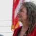 A middle-aged to older woman is photographed in profile view, gazing upward or into the distance. She is wearing a red jacket with a small pin or brooch on the lapel, with an American flag visible in the background. The image appears to be taken at an outdoor public or civic event.