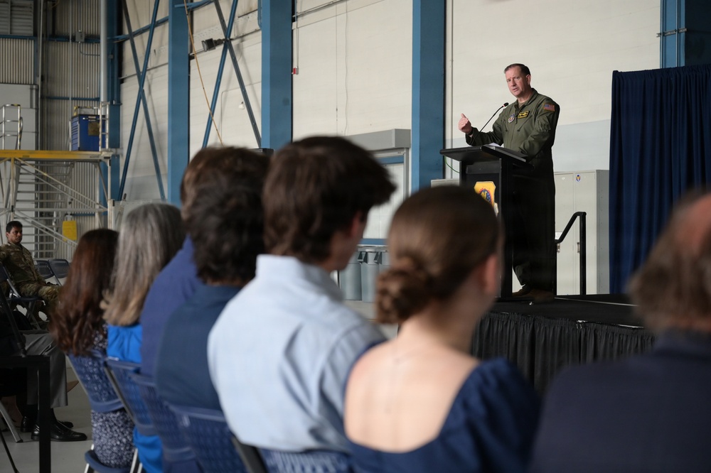 A military officer in a green flight suit is delivering a speech at a podium on a raised stage inside what appears to be an aircraft hangar. An audience of civilians and at least one uniformed military personnel are seated in chairs facing the speaker. The setting suggests a formal military ceremony or briefing event.