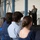 A military officer in a green flight suit is delivering a speech at a podium on a raised stage inside what appears to be an aircraft hangar. An audience of civilians and at least one uniformed military personnel are seated in chairs facing the speaker. The setting suggests a formal military ceremony or briefing event.
