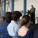 A military officer in a green flight suit is delivering a speech at a podium on a raised stage inside what appears to be an aircraft hangar. An audience of civilians and at least one uniformed military personnel are seated in chairs facing the speaker. The setting suggests a formal military ceremony or briefing event.
