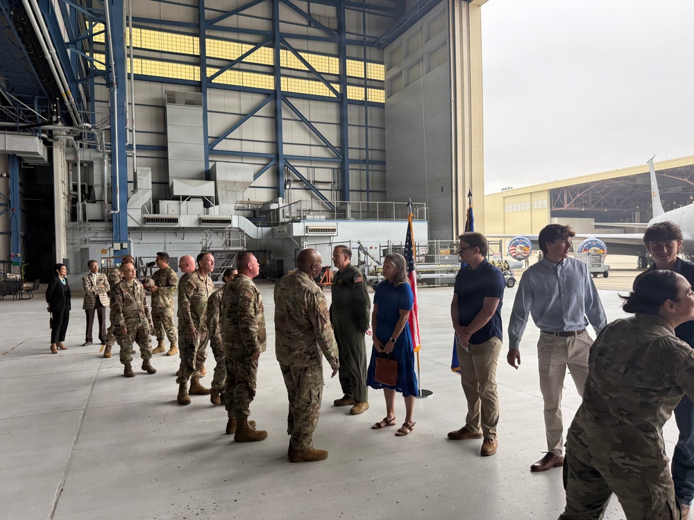 A military ceremony or reception is taking place inside a large aircraft hangar. A line of uniformed military personnel in camouflage (OCP) uniforms appears to be greeting or being introduced to civilians, including a woman in a blue dress and several young men in casual attire. An American flag and what appears to be a unit flag are visible in the background near the hangar opening.