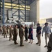 A military ceremony or reception is taking place inside a large aircraft hangar. A line of uniformed military personnel in camouflage (OCP) uniforms appears to be greeting or being introduced to civilians, including a woman in a blue dress and several young men in casual attire. An American flag and what appears to be a unit flag are visible in the background near the hangar opening.