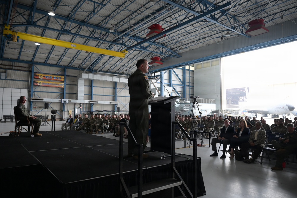 A military officer in flight suit is delivering a speech at a podium on a raised stage inside a large aircraft hangar. A large audience of military personnel and civilians is seated in rows facing the speaker. A military aircraft, likely a KC-135 or similar tanker, is visible through the open hangar doors in the background.