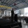 A military officer in flight suit is delivering a speech at a podium on a raised stage inside a large aircraft hangar. A large audience of military personnel and civilians is seated in rows facing the speaker. A military aircraft, likely a KC-135 or similar tanker, is visible through the open hangar doors in the background.