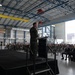 A military officer in flight suit is delivering a speech at a podium on a raised stage inside a large aircraft hangar. A large audience of military personnel and civilians is seated in rows facing the speaker. A military aircraft, likely a KC-135 or similar tanker, is visible through the open hangar doors in the background.