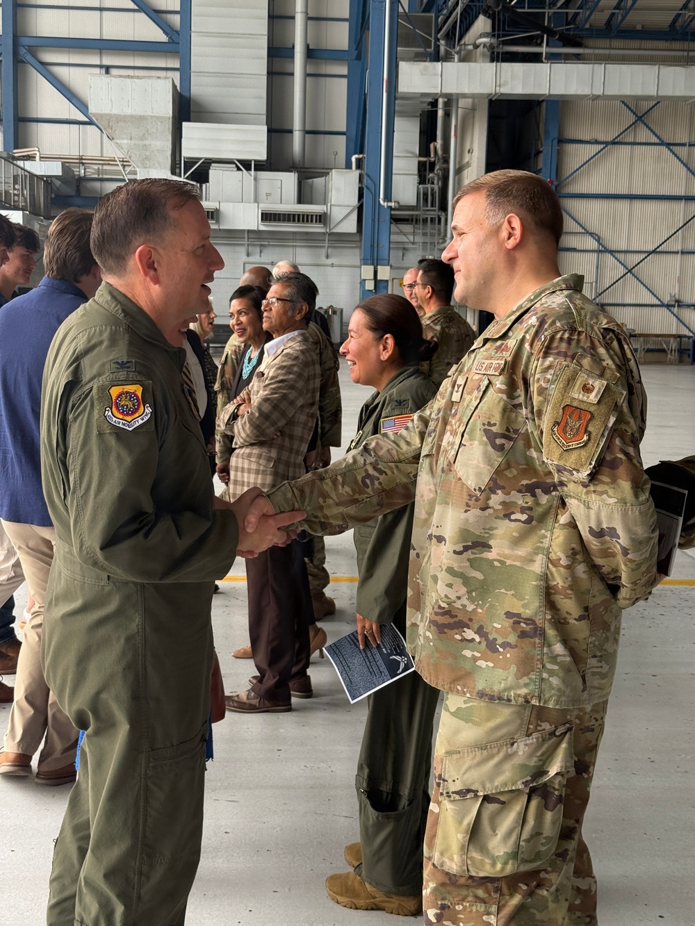 Two U.S. Air Force personnel are shaking hands inside what appears to be a military aircraft hangar. The individual on the left is wearing an olive green flight suit with a unit patch, while the individual on the right is wearing OCP (Operational Camouflage Pattern) uniform with a U.S. Air Force tape visible. Several other military and civilian personnel are visible in the background engaged in conversation.