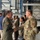 Two U.S. Air Force personnel are shaking hands inside what appears to be a military aircraft hangar. The individual on the left is wearing an olive green flight suit with a unit patch, while the individual on the right is wearing OCP (Operational Camouflage Pattern) uniform with a U.S. Air Force tape visible. Several other military and civilian personnel are visible in the background engaged in conversation.