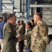 Two U.S. Air Force personnel are shaking hands inside what appears to be a military aircraft hangar. The individual on the left is wearing an olive green flight suit with a unit patch, while the individual on the right is wearing OCP (Operational Camouflage Pattern) uniform with a U.S. Air Force tape visible. Several other military and civilian personnel are visible in the background engaged in conversation.