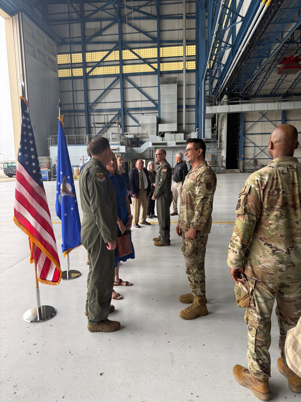A military ceremony or reception is taking place inside a large aircraft hangar. Personnel in US Air Force flight suits and OCP (Operational Camouflage Pattern) uniforms are interacting with civilian guests in a formal line or receiving formation. The US flag and an Air Force organizational flag are displayed on stands to the left.