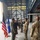 A military ceremony or reception is taking place inside a large aircraft hangar. Personnel in US Air Force flight suits and OCP (Operational Camouflage Pattern) uniforms are interacting with civilian guests in a formal line or receiving formation. The US flag and an Air Force organizational flag are displayed on stands to the left.