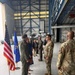 A military ceremony or reception is taking place inside a large aircraft hangar. Personnel in US Air Force flight suits and OCP (Operational Camouflage Pattern) uniforms are interacting with civilian guests in a formal line or receiving formation. The US flag and an Air Force organizational flag are displayed on stands to the left.
