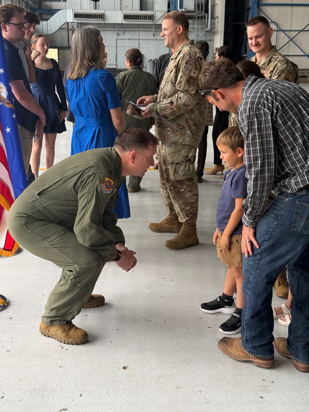 A military ceremony taking place inside what appears to be an aircraft hangar. A uniformed airman in an olive green flight suit is crouching down to interact with a young child standing next to an adult male in civilian clothing. Other military personnel in camouflage uniforms and civilians are visible in the background near an American flag.