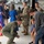 A military ceremony taking place inside what appears to be an aircraft hangar. A uniformed airman in an olive green flight suit is crouching down to interact with a young child standing next to an adult male in civilian clothing. Other military personnel in camouflage uniforms and civilians are visible in the background near an American flag.