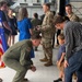 A military ceremony taking place inside what appears to be an aircraft hangar. A uniformed airman in an olive green flight suit is crouching down to interact with a young child standing next to an adult male in civilian clothing. Other military personnel in camouflage uniforms and civilians are visible in the background near an American flag.