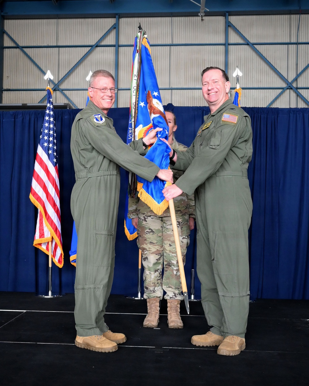 Two U.S. Air Force officers in olive green flight suits are participating in a formal change of command ceremony, jointly holding a blue unit guidon with gold fringe. The ceremony is taking place in what appears to be a military hangar decorated with blue draping, with an American flag and Air Force flag visible in the background. A third service member in camouflage uniform stands behind them as a guidon bearer.