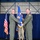 Two U.S. Air Force officers in olive green flight suits are participating in a formal change of command ceremony, jointly holding a blue unit guidon with gold fringe. The ceremony is taking place in what appears to be a military hangar decorated with blue draping, with an American flag and Air Force flag visible in the background. A third service member in camouflage uniform stands behind them as a guidon bearer.