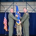Two U.S. Air Force officers in olive green flight suits are participating in a formal change of command ceremony, jointly holding a blue unit guidon with gold fringe. The ceremony is taking place in what appears to be a military hangar decorated with blue draping, with an American flag and Air Force flag visible in the background. A third service member in camouflage uniform stands behind them as a guidon bearer.
