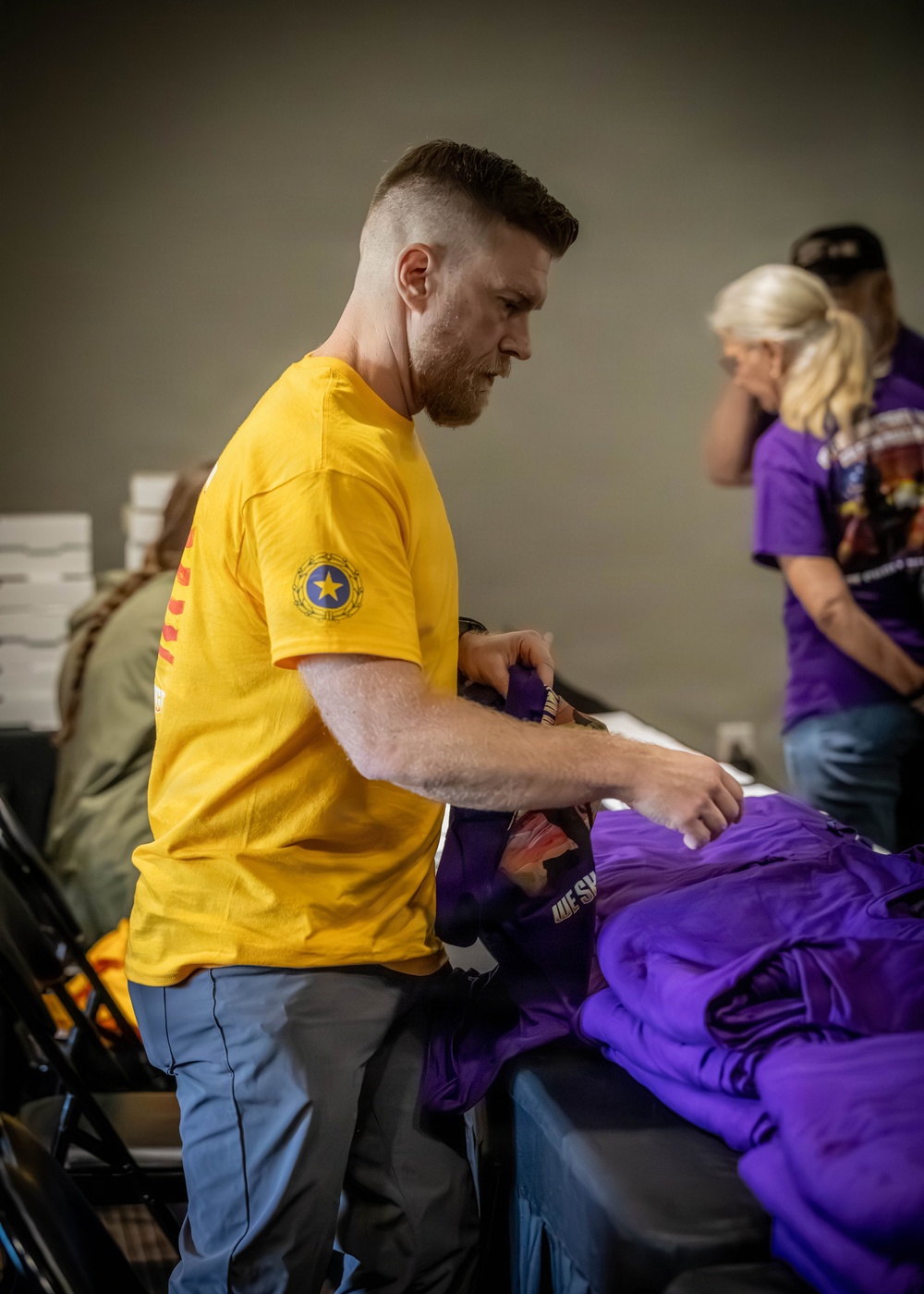 A man wearing a yellow t-shirt with a star emblem on the sleeve is sorting or folding purple shirts at a table. The setting appears to be an indoor event or volunteer activity, with other individuals visible in the background also wearing purple shirts. The scene suggests an organized distribution or preparation effort.