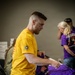 A man wearing a yellow t-shirt with a star emblem on the sleeve is sorting or folding purple shirts at a table. The setting appears to be an indoor event or volunteer activity, with other individuals visible in the background also wearing purple shirts. The scene suggests an organized distribution or preparation effort.