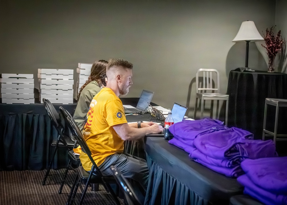 Two individuals are seated at a draped table in what appears to be an event registration or operations area, working on laptops. The man in the foreground wears a yellow t-shirt with a star emblem and is actively typing, while a woman behind him wears a headset. The setting includes stacked pizza boxes, folded purple shirts, a decorative lamp, and a metal chair, suggesting a behind-the-scenes event coordination space.