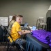 Two individuals are seated at a draped table in what appears to be an event registration or operations area, working on laptops. The man in the foreground wears a yellow t-shirt with a star emblem and is actively typing, while a woman behind him wears a headset. The setting includes stacked pizza boxes, folded purple shirts, a decorative lamp, and a metal chair, suggesting a behind-the-scenes event coordination space.