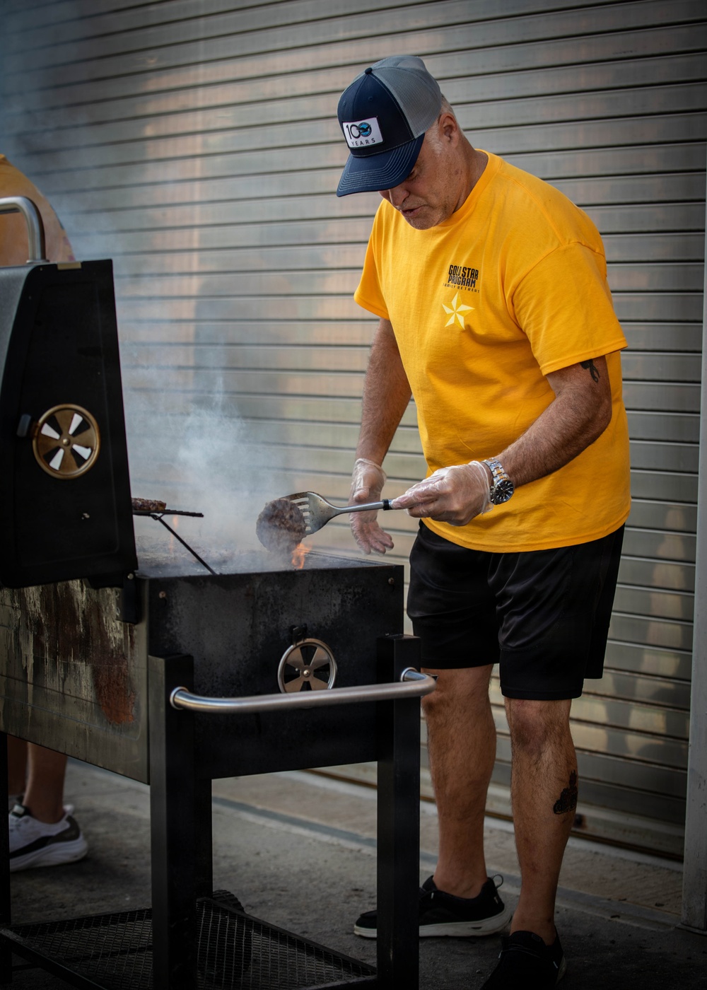 A middle-aged man wearing a yellow 'Gold Star Program' t-shirt and a navy/gray '100 Years' trucker cap is grilling meat on a charcoal grill outdoors. He is using a spatula and wearing clear plastic gloves while tending to food over visible flames and smoke. The setting appears to be an outdoor area with a corrugated metal wall in the background.