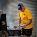 A middle-aged man wearing a yellow 'Gold Star Program' t-shirt and a navy/gray '100 Years' trucker cap is grilling meat on a charcoal grill outdoors. He is using a spatula and wearing clear plastic gloves while tending to food over visible flames and smoke. The setting appears to be an outdoor area with a corrugated metal wall in the background.