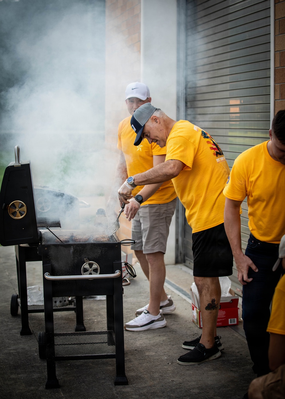 A group of men wearing matching yellow t-shirts are gathered outdoors around a large black charcoal/smoker grill. One man, wearing a gray cap and watch, is actively tending to the grill with tongs while smoke billows heavily around them. The setting appears to be a community or organized group event, likely a barbecue or cookout near a building with a roller shutter door.