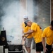 A group of men wearing matching yellow t-shirts are gathered outdoors around a large black charcoal/smoker grill. One man, wearing a gray cap and watch, is actively tending to the grill with tongs while smoke billows heavily around them. The setting appears to be a community or organized group event, likely a barbecue or cookout near a building with a roller shutter door.