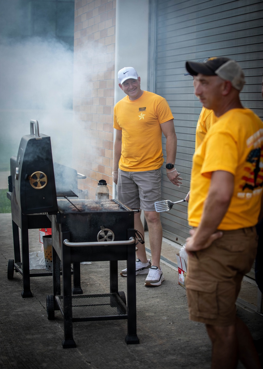 Two men wearing matching yellow 'Gold Star' t-shirts are grilling outdoors next to a large black charcoal smoker/grill. The man in the background is smiling and wearing a white Nike cap, while the man in the foreground holds a metal spatula. Significant smoke is billowing from the grill, suggesting active cooking.