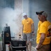Two men wearing matching yellow 'Gold Star' t-shirts are grilling outdoors next to a large black charcoal smoker/grill. The man in the background is smiling and wearing a white Nike cap, while the man in the foreground holds a metal spatula. Significant smoke is billowing from the grill, suggesting active cooking.