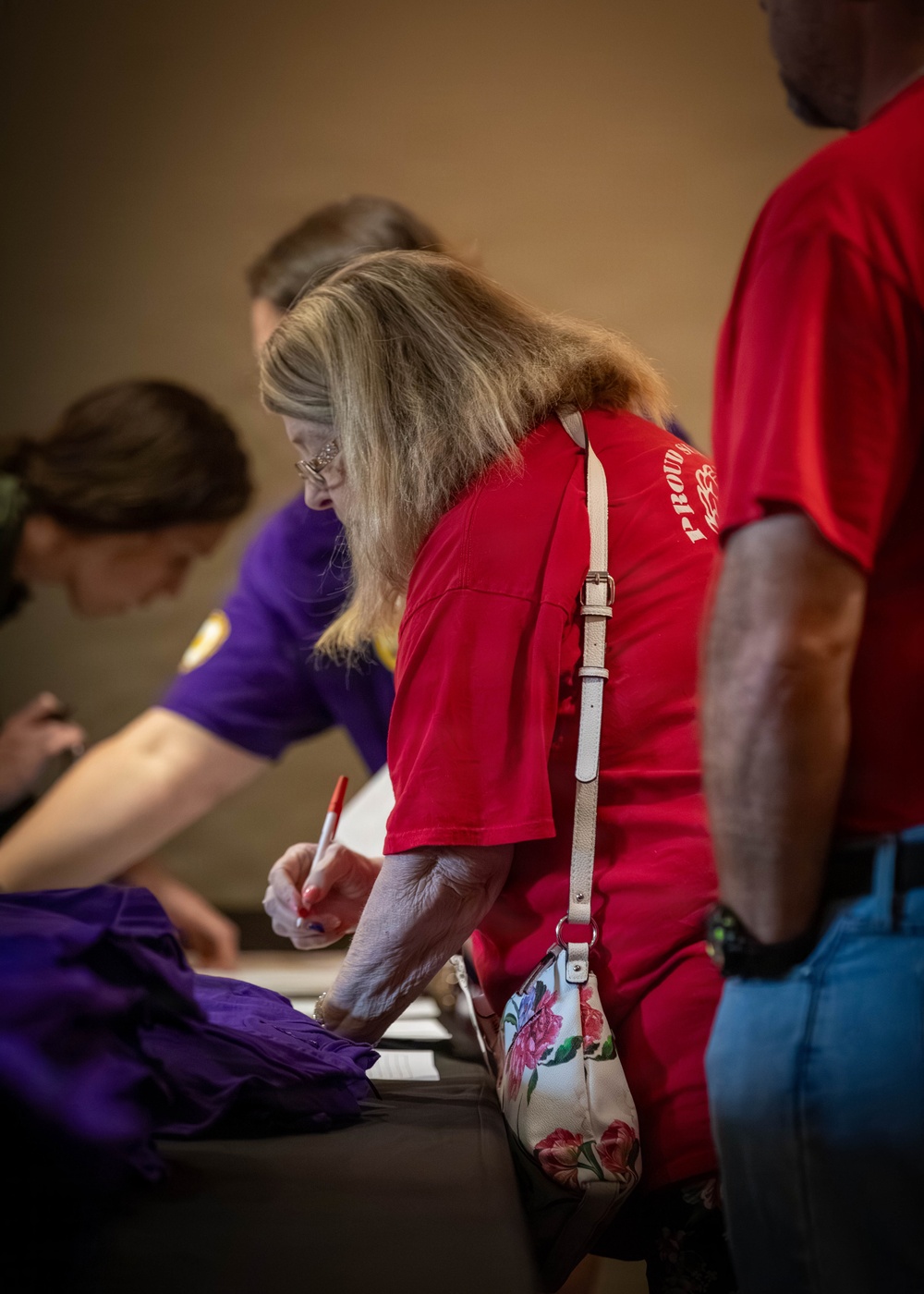 The image shows several people gathered around a table in what appears to be a registration or sign-in event. A woman in a red shirt with blonde hair and glasses is leaning over the table, writing with a red pen on a document. She carries a white floral-patterned crossbody bag.