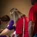 The image shows several people gathered around a table in what appears to be a registration or sign-in event. A woman in a red shirt with blonde hair and glasses is leaning over the table, writing with a red pen on a document. She carries a white floral-patterned crossbody bag.