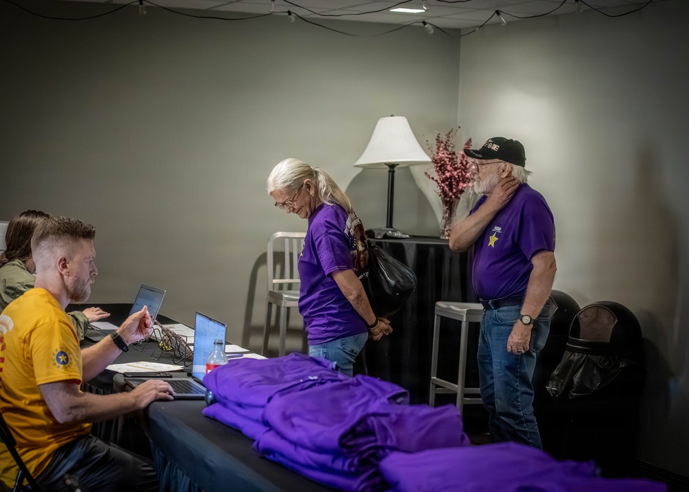 A registration or check-in table scene where two older adults wearing matching purple shirts with a gold star emblem are being assisted by a younger man in a yellow shirt working on a laptop. Purple folded shirts are stacked on the table, suggesting a distribution or sign-in event. The setting appears to be an indoor venue with temporary lighting and décor.