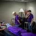 A registration or check-in table scene where two older adults wearing matching purple shirts with a gold star emblem are being assisted by a younger man in a yellow shirt working on a laptop. Purple folded shirts are stacked on the table, suggesting a distribution or sign-in event. The setting appears to be an indoor venue with temporary lighting and décor.