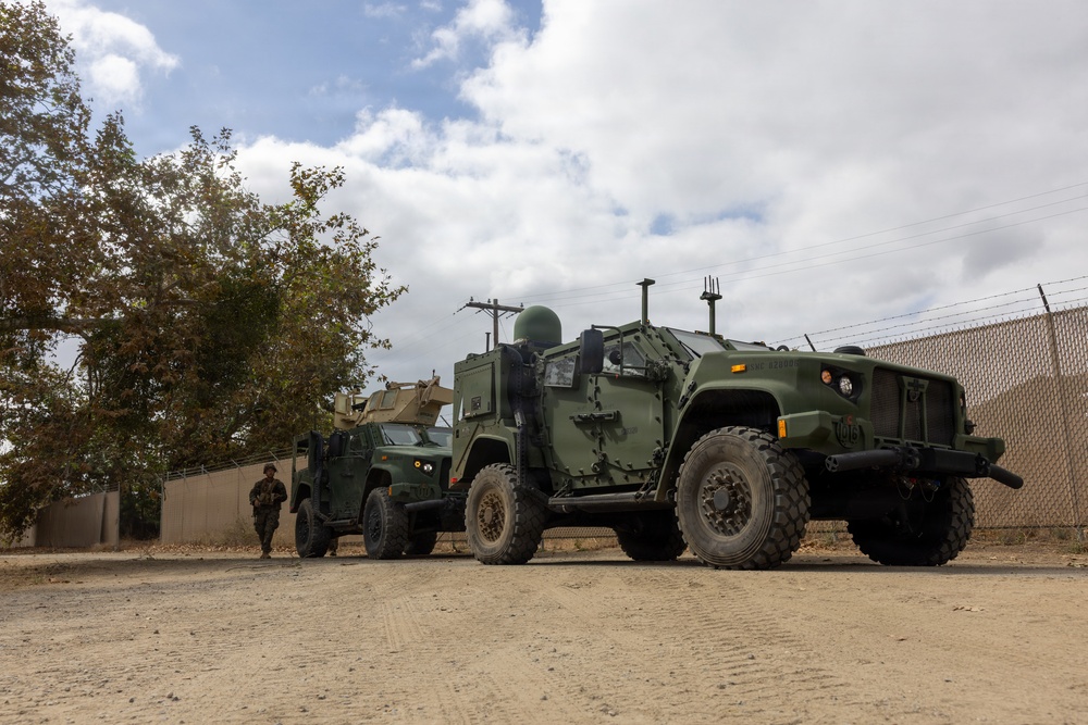 The image shows two military tactical vehicles parked on a dirt/gravel surface near a perimeter fence with barbed wire. At least one uniformed soldier is visible standing between the vehicles. The setting appears to be a military installation or secured compound.