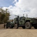 The image shows two military tactical vehicles parked on a dirt/gravel surface near a perimeter fence with barbed wire. At least one uniformed soldier is visible standing between the vehicles. The setting appears to be a military installation or secured compound.