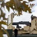 A soldier is positioned in the turret hatch of a tan/desert-colored armored military vehicle, manning a mounted machine gun. The image is taken from a low angle through out-of-focus tree branches, giving a partially obscured view of the scene. The vehicle appears to be a modern U.S. military armored platform with ballistic glass panels and external armor plating.
