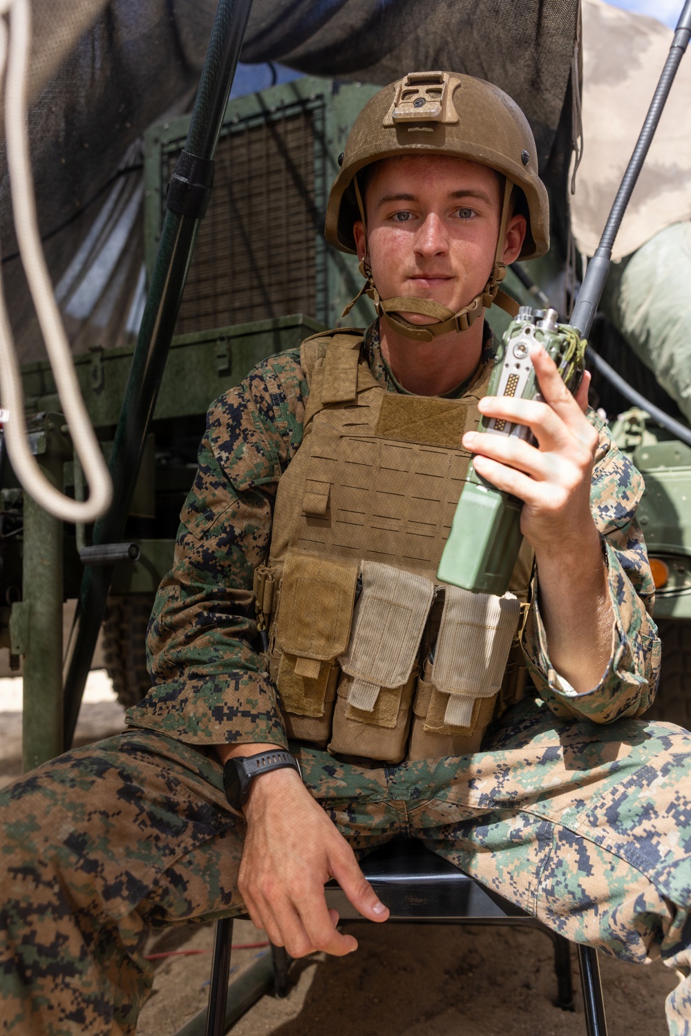 A young male service member in U.S. Marine Corps MARPAT digital camouflage uniform is seated near a military vehicle, holding a handheld tactical radio. He is wearing a plate carrier vest, a combat helmet with a NVG mount bracket, and a chin strap. The setting appears to be an outdoor field environment, likely during a training exercise or operational deployment.