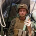 A young male service member in U.S. Marine Corps MARPAT digital camouflage uniform is seated near a military vehicle, holding a handheld tactical radio. He is wearing a plate carrier vest, a combat helmet with a NVG mount bracket, and a chin strap. The setting appears to be an outdoor field environment, likely during a training exercise or operational deployment.