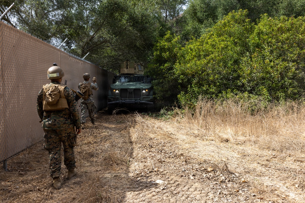 Two military personnel in camouflage uniforms are walking along a chain-link fence line toward a military armored vehicle partially obscured by dense vegetation. The vehicle appears to be a Mine-Resistant Ambush Protected (MRAP) or similar tactical vehicle with its headlights illuminated. The setting appears to be a dry, brushy terrain consistent with a training environment or operational area.