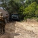Two military personnel in camouflage uniforms are walking along a chain-link fence line toward a military armored vehicle partially obscured by dense vegetation. The vehicle appears to be a Mine-Resistant Ambush Protected (MRAP) or similar tactical vehicle with its headlights illuminated. The setting appears to be a dry, brushy terrain consistent with a training environment or operational area.