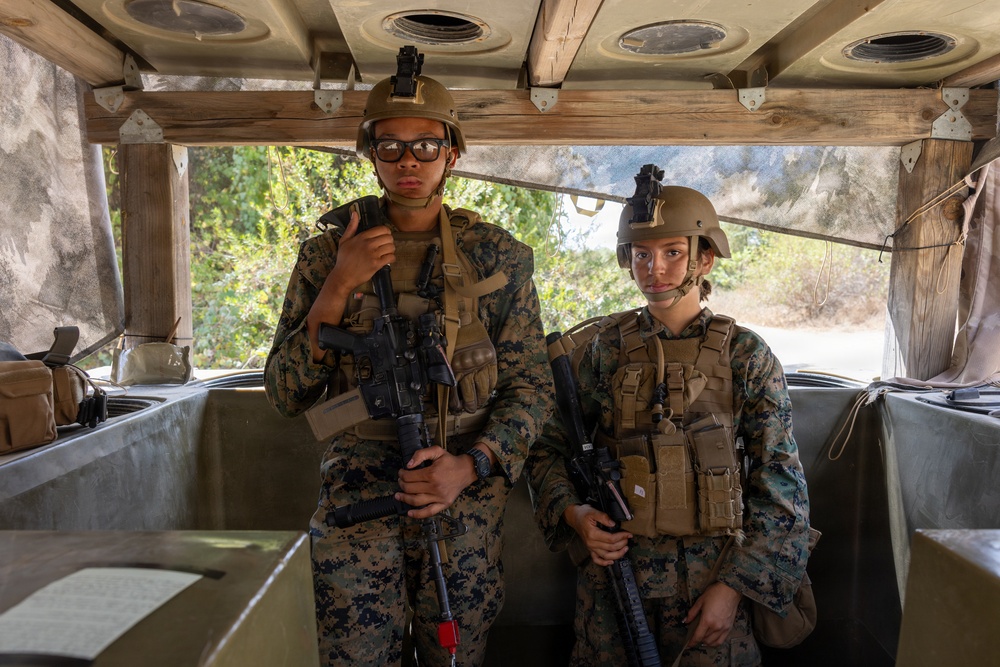 Two military personnel in full combat gear are positioned inside what appears to be a military vehicle, likely a HMMWV (Humvee) or similar tactical transport. Both individuals are wearing MARPAT (Marine pattern) camouflage uniforms, tactical vests, and helmets with mounted accessories, and are holding rifles. The setting suggests a training exercise or operational deployment.