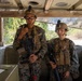 Two military personnel in full combat gear are positioned inside what appears to be a military vehicle, likely a HMMWV (Humvee) or similar tactical transport. Both individuals are wearing MARPAT (Marine pattern) camouflage uniforms, tactical vests, and helmets with mounted accessories, and are holding rifles. The setting suggests a training exercise or operational deployment.