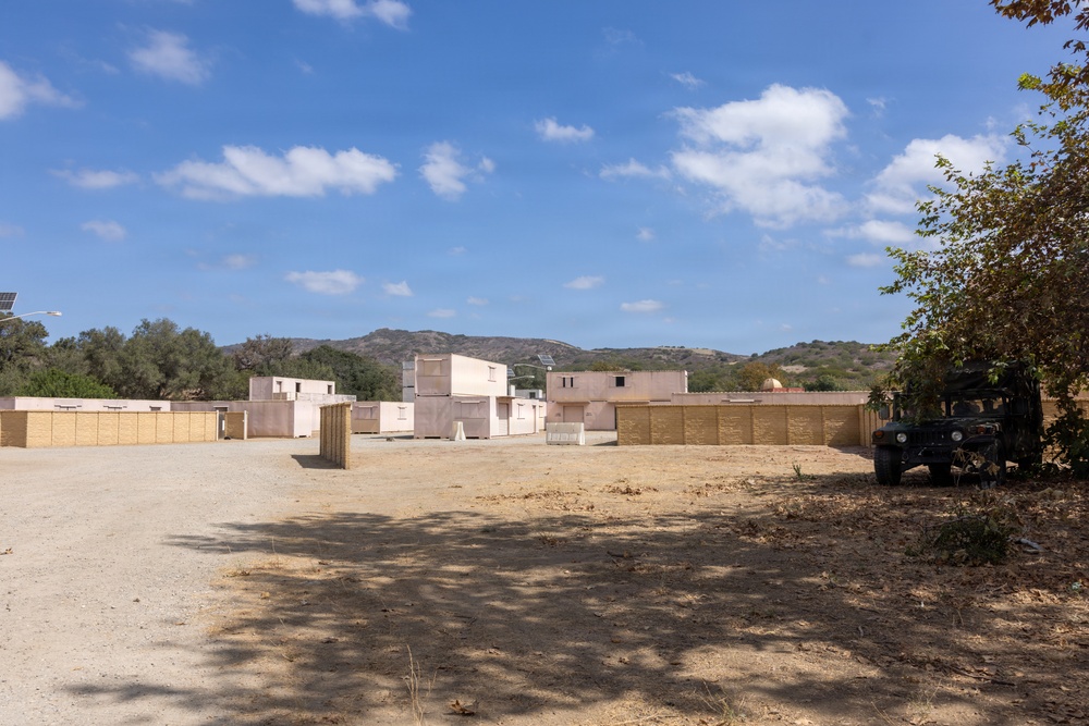 The image shows what appears to be a military urban training facility (MOUT - Military Operations on Urban Terrain) with mock buildings constructed to simulate a Middle Eastern or desert village environment. The structures are simple, flat-roofed, and tan/beige in color, arranged to replicate an urban layout. A military Humvee is parked on the right side, and solar panels are visible, suggesting an active training installation.