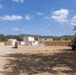 The image shows what appears to be a military urban training facility (MOUT - Military Operations on Urban Terrain) with mock buildings constructed to simulate a Middle Eastern or desert village environment. The structures are simple, flat-roofed, and tan/beige in color, arranged to replicate an urban layout. A military Humvee is parked on the right side, and solar panels are visible, suggesting an active training installation.